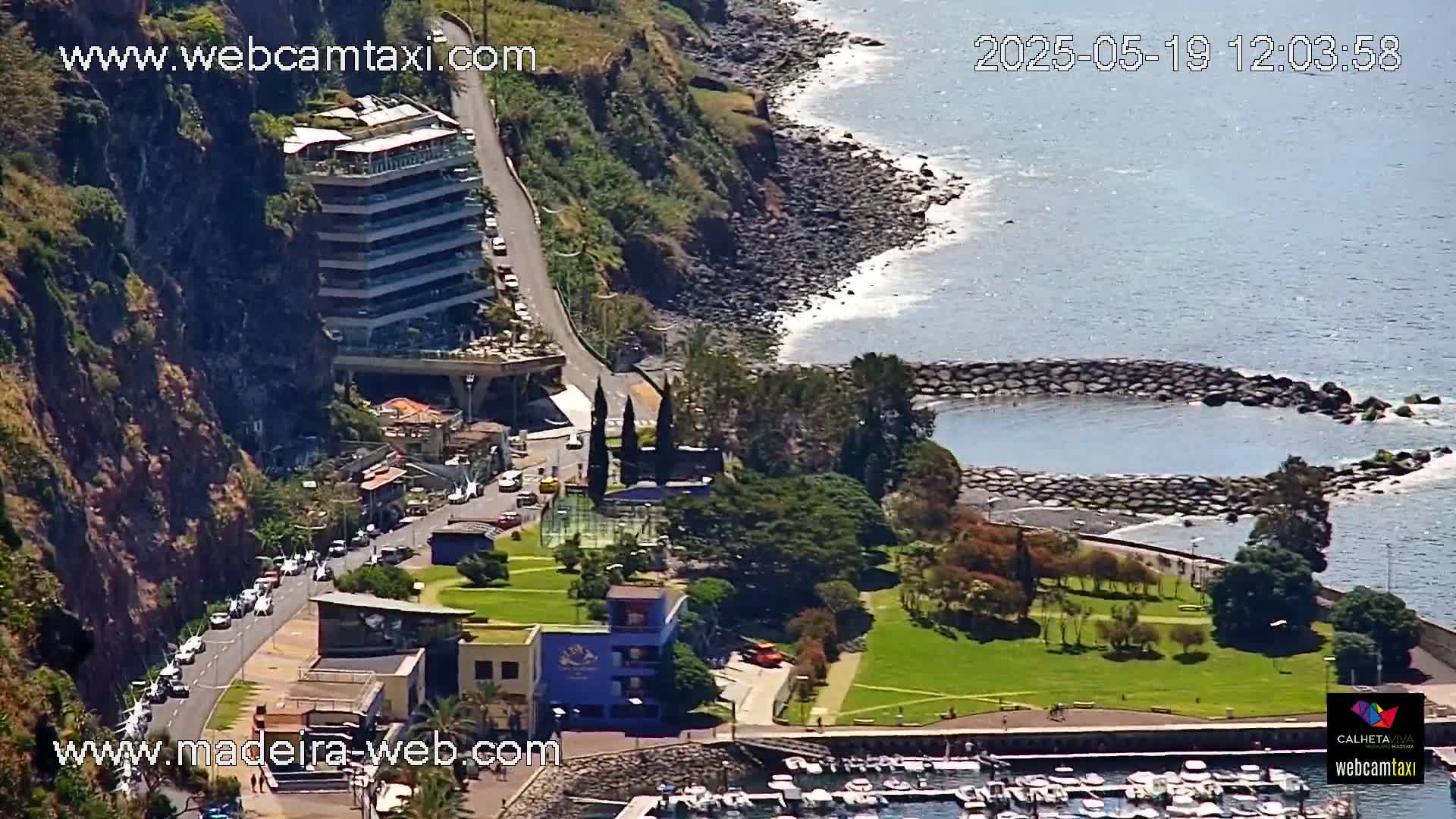A coastal town scene on a sunny day shows a multi-story building on a cliff overlooking a small marina and a grassy park area next to a rocky shoreline and calm ocean water.