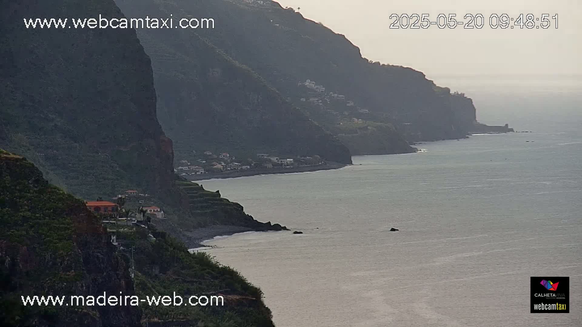 A hazy coastal view shows dark green cliffs descending to a calm ocean with a small village nestled along the shore.