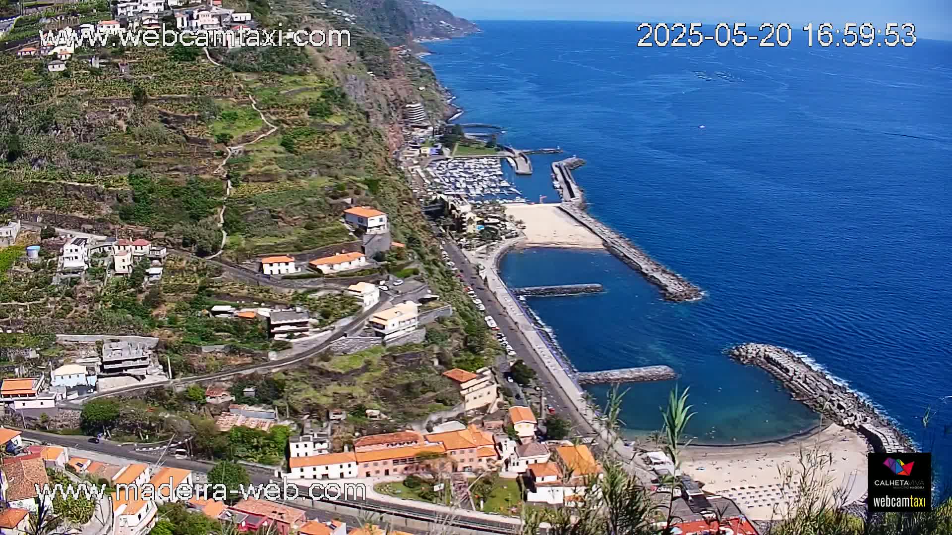 A coastal town nestled on a steep hillside overlooking a marina and a small, sandy beach on a sunny day.
