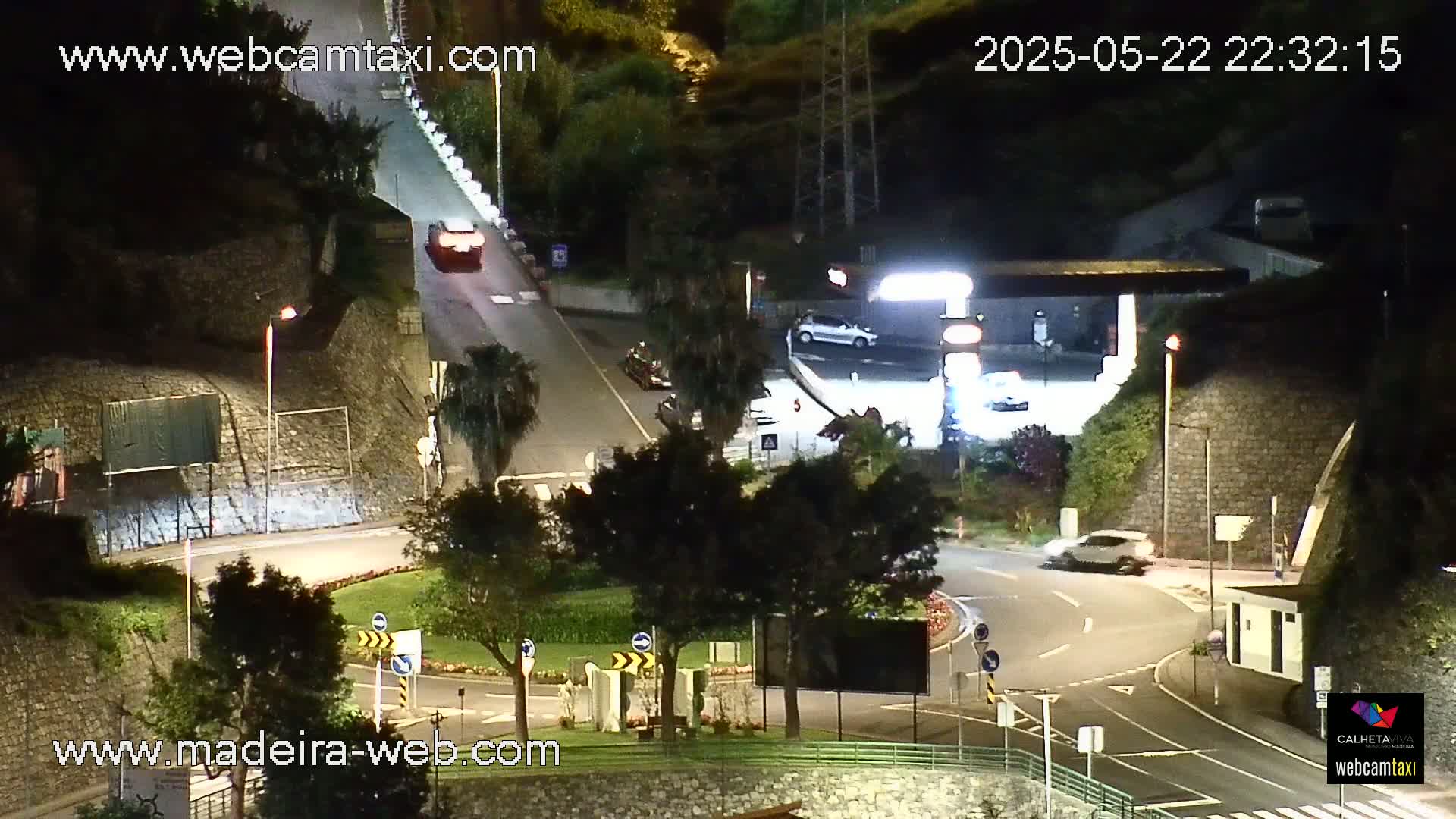 A nighttime, overhead view of a roadway intersection with several cars, including one at a gas station, and lush green landscaping illuminated by streetlights.