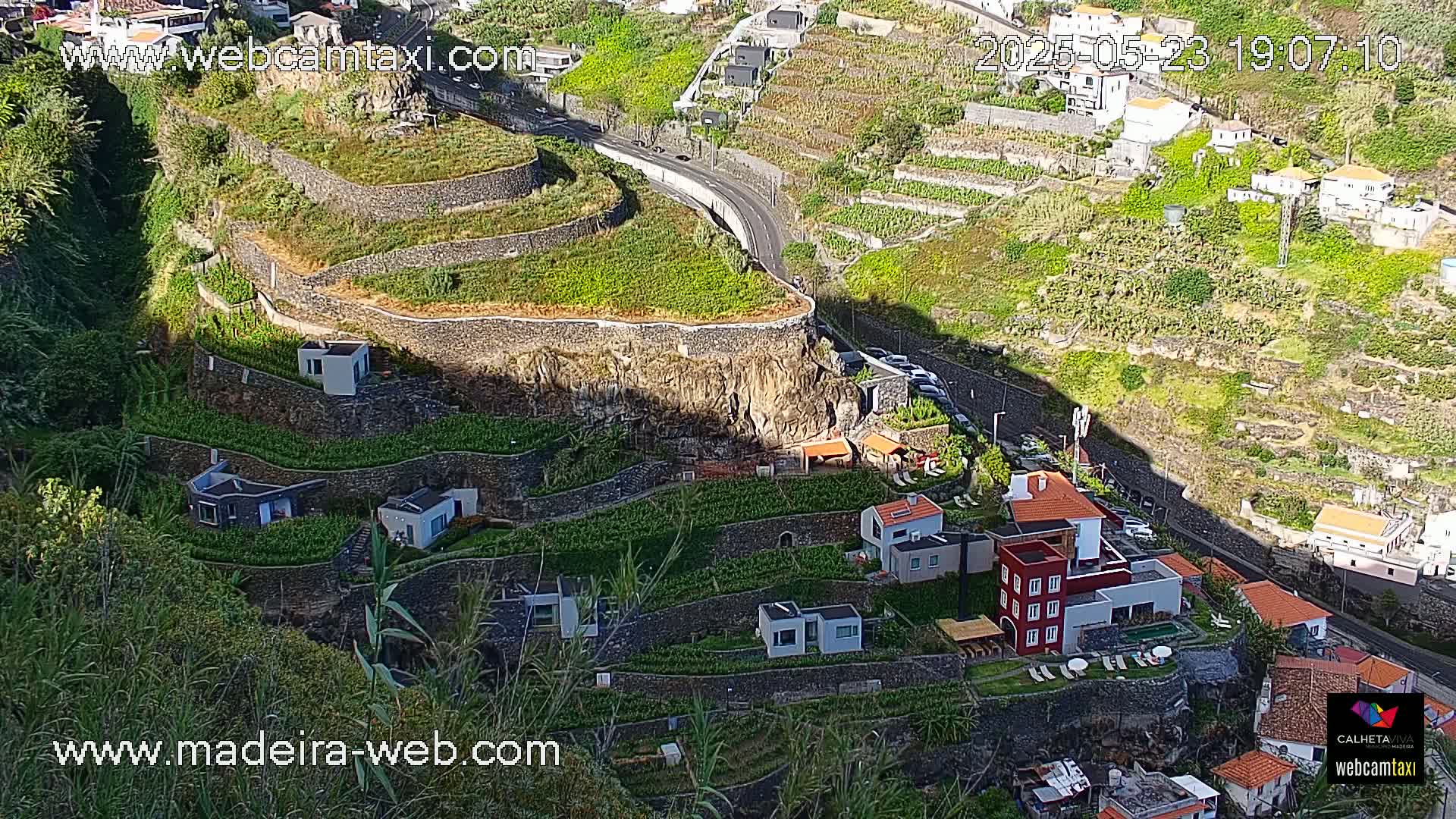 A sunny, high-angle view shows terraced farmland with several houses built into the hillside, alongside a winding road.