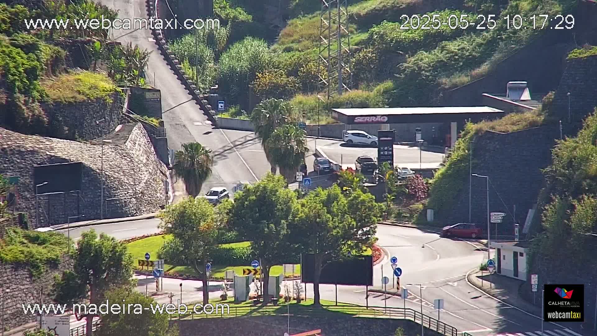 A sunny day reveals a roadway curving around a roundabout near a gas station, bordered by stone walls and lush greenery.