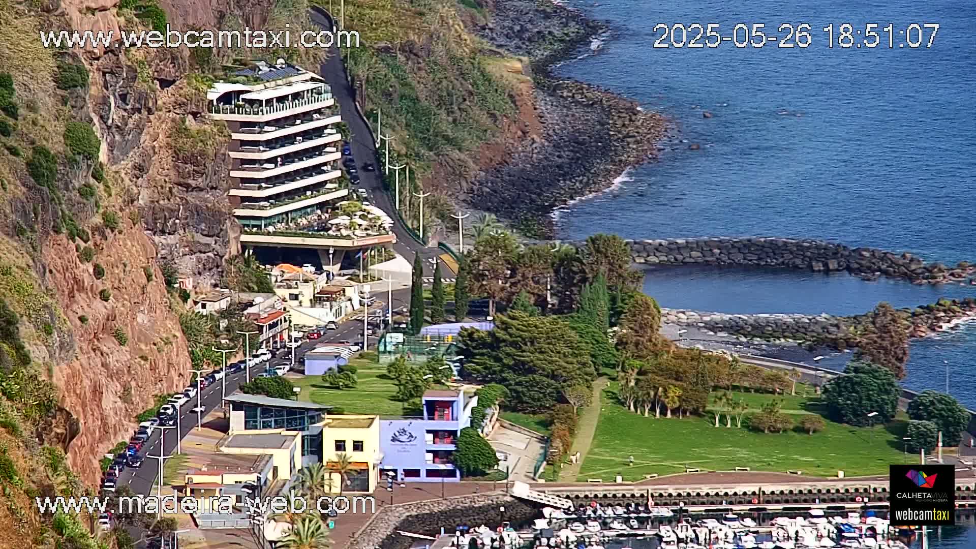 A seaside town with a multi-story building built into a cliffside overlooks a calm, blue ocean and a marina filled with boats, all under a sunny sky.