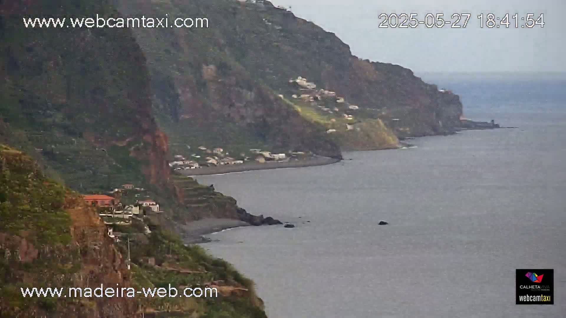 A dark, overcast day reveals a rocky, green coastline with a small village nestled into the cliffside overlooking a calm, grey ocean.