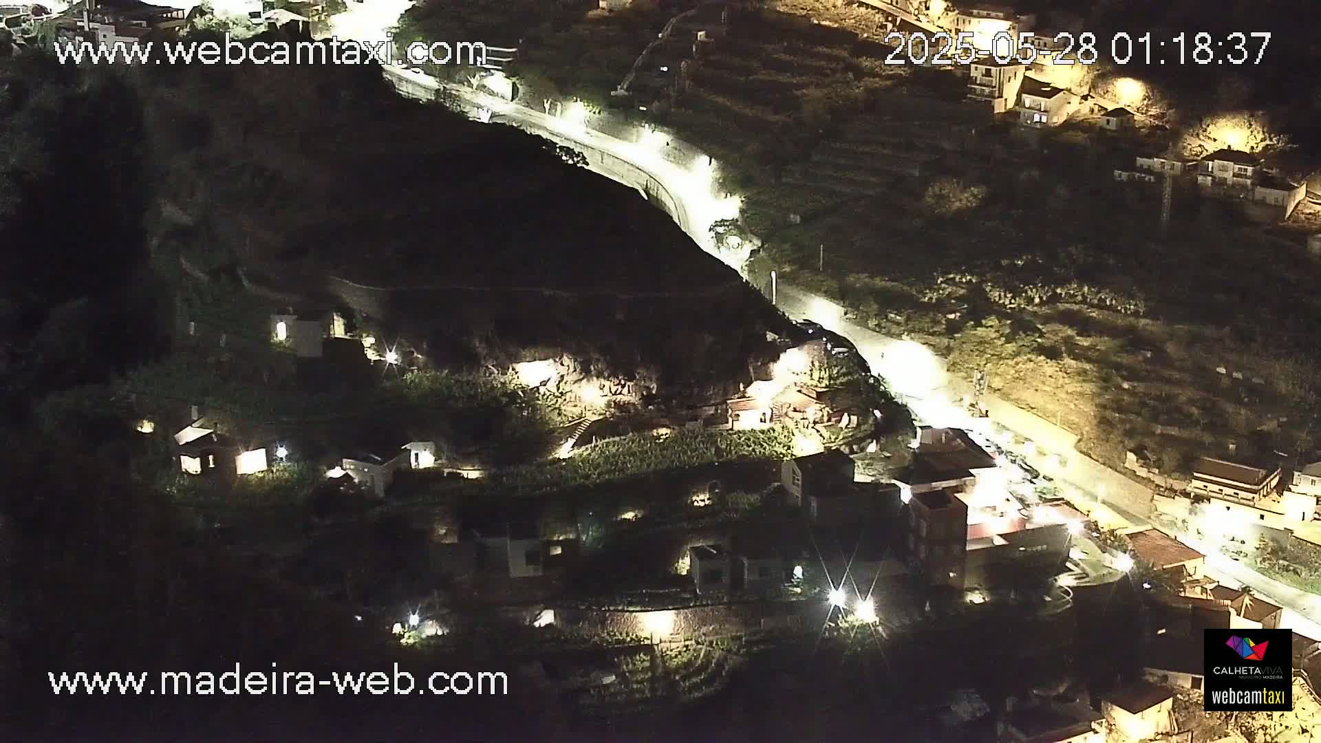 A nighttime aerial view of a hillside village with illuminated buildings and a winding road, under clear skies.