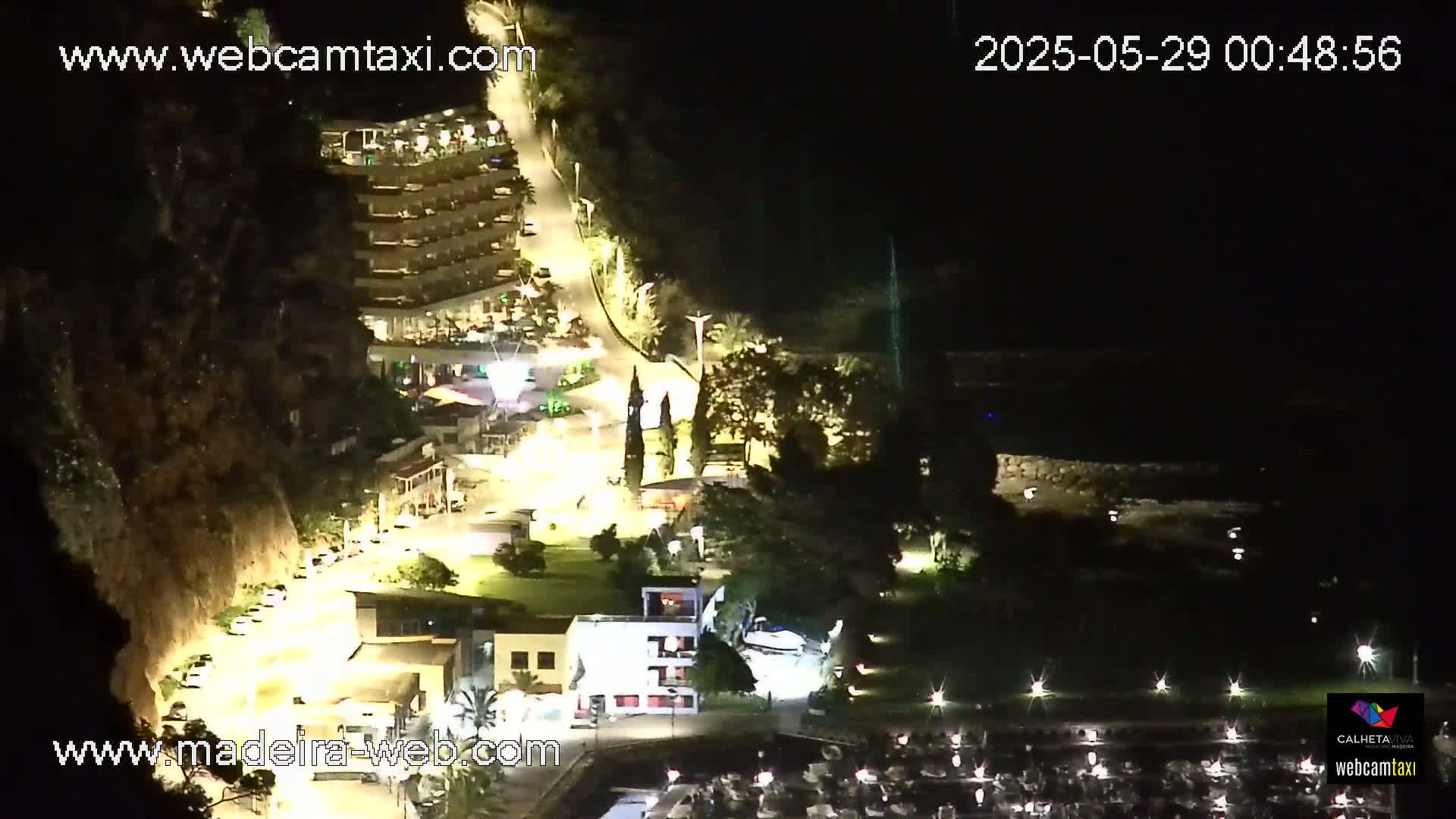 A nighttime aerial view of a coastal town shows a marina filled with boats, buildings illuminated along a winding road, and dark cliffs rising behind them.