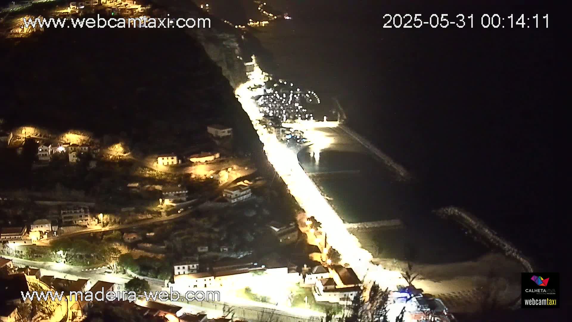 A nighttime, high-angle view shows a brightly lit coastal road winding through a village built into a dark hillside, alongside a dark body of water.