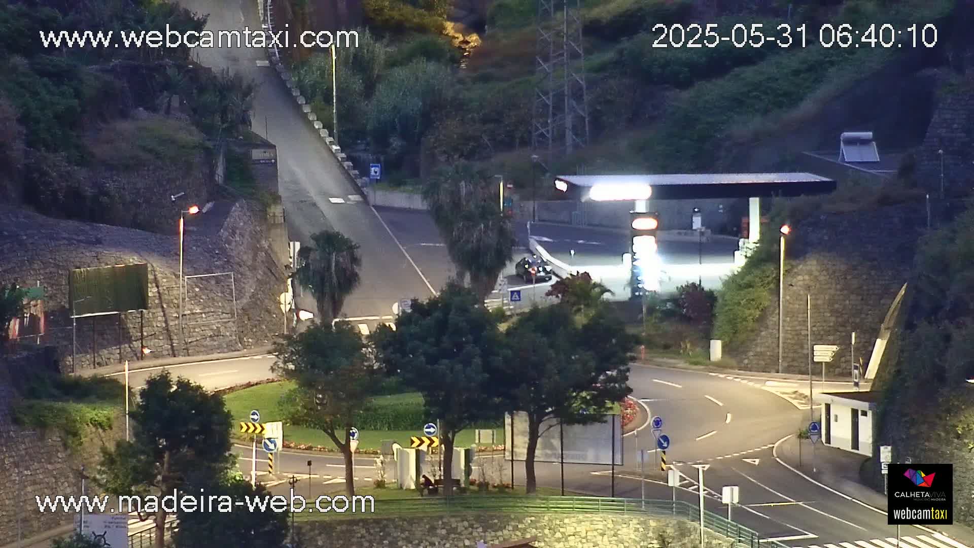 An elevated view at night shows a roadway curving around a small traffic circle with sparse landscaping, under partly cloudy skies.