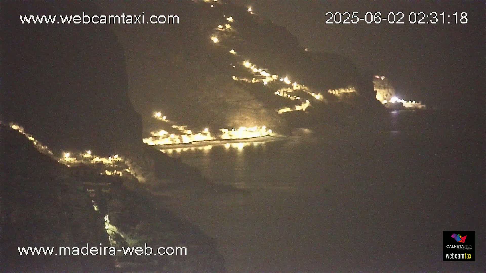 A nighttime view of a coastal town illuminated by lights, nestled against dark hillsides and overlooking a calm body of water.