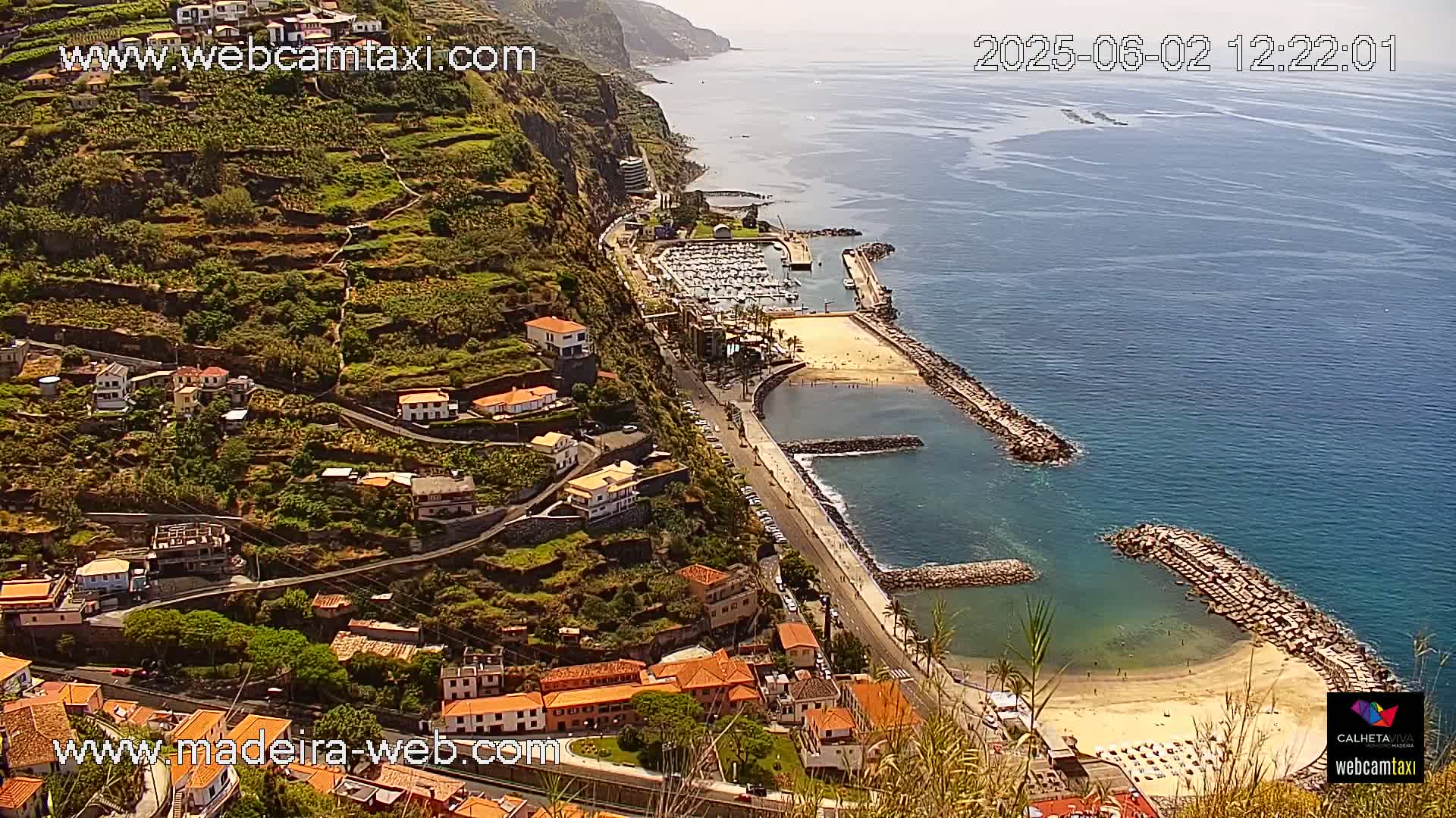 A coastal town nestled on a hillside overlooks a calm ocean harbor with a sandy beach and a marina on a sunny day.