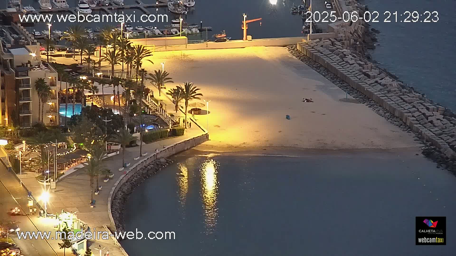 A nighttime aerial view of a sandy beach illuminated by artificial lights, adjacent to a building with a pool, a paved walkway, and a harbor in the background.