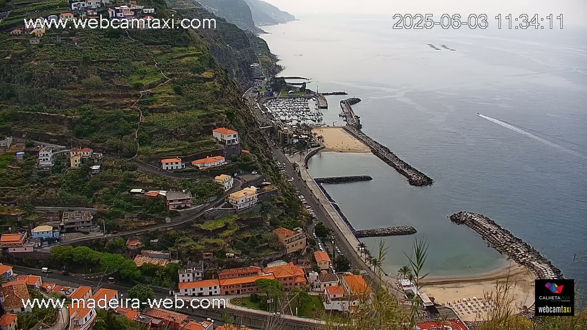 An aerial view shows a coastal town nestled on a steep hillside overlooking a calm ocean with a marina and small sandy beach.