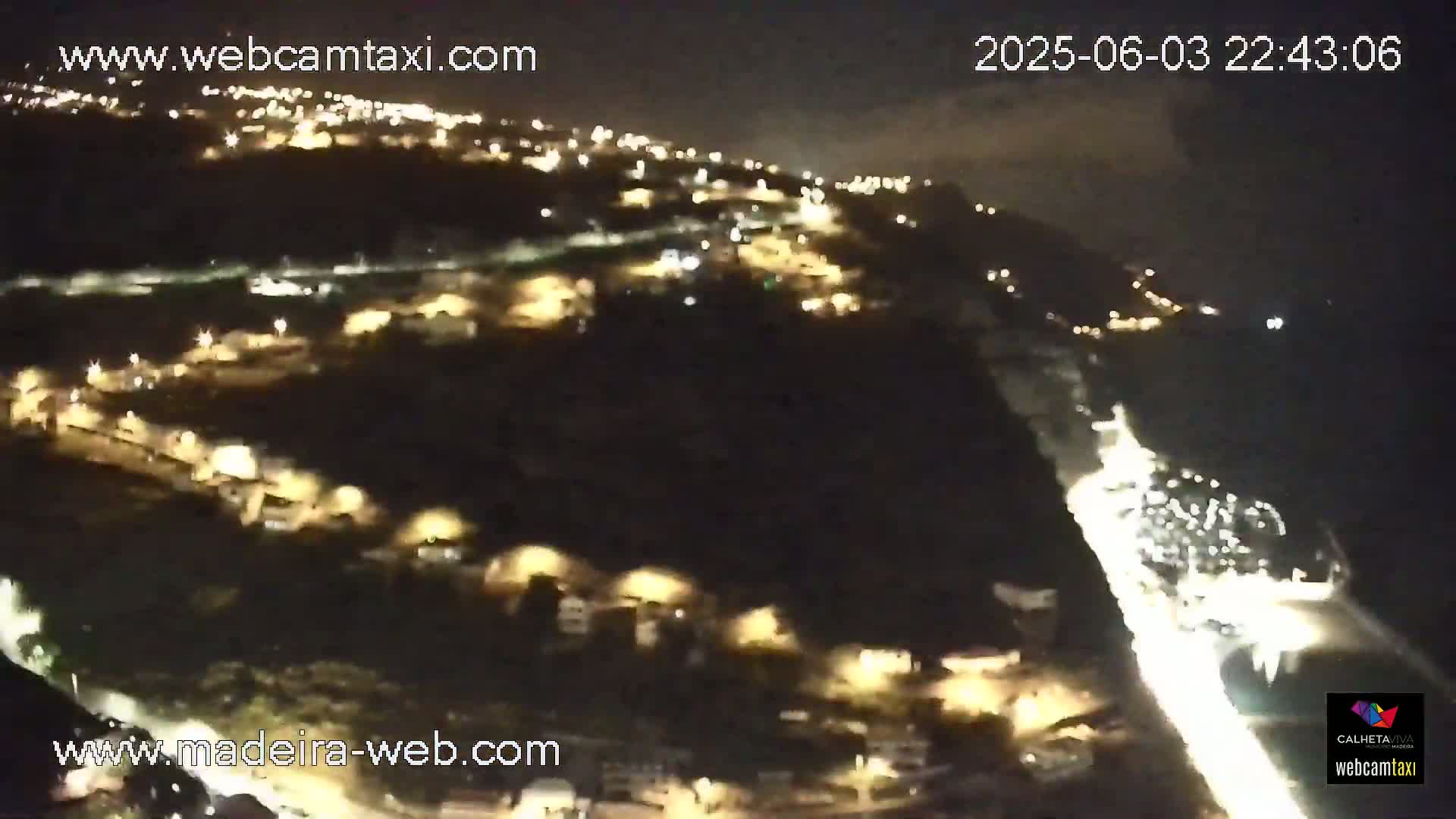 A nighttime, high-angle view shows a coastal town illuminated with many small lights, alongside a bright roadway curving along the water's edge.