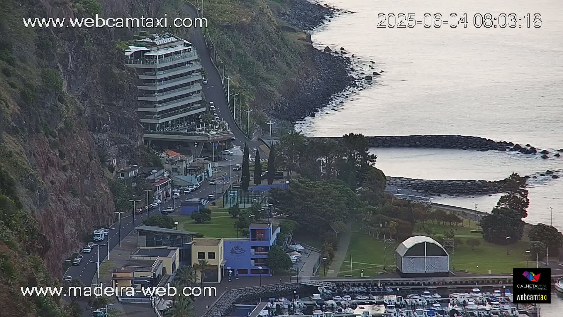 A coastal scene shows a multi-story building perched on a cliff overlooking a calm ocean with a marina and green parkland below.