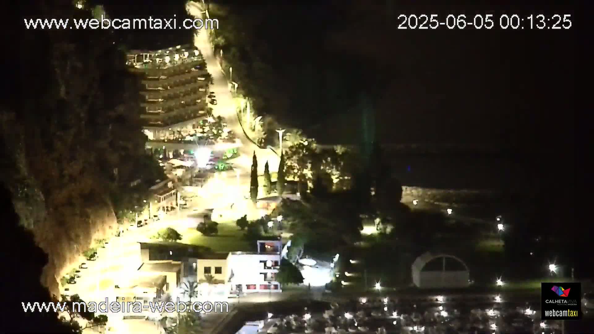A nighttime aerial view shows a coastal area with buildings, roads, and a marina, illuminated by artificial lights.