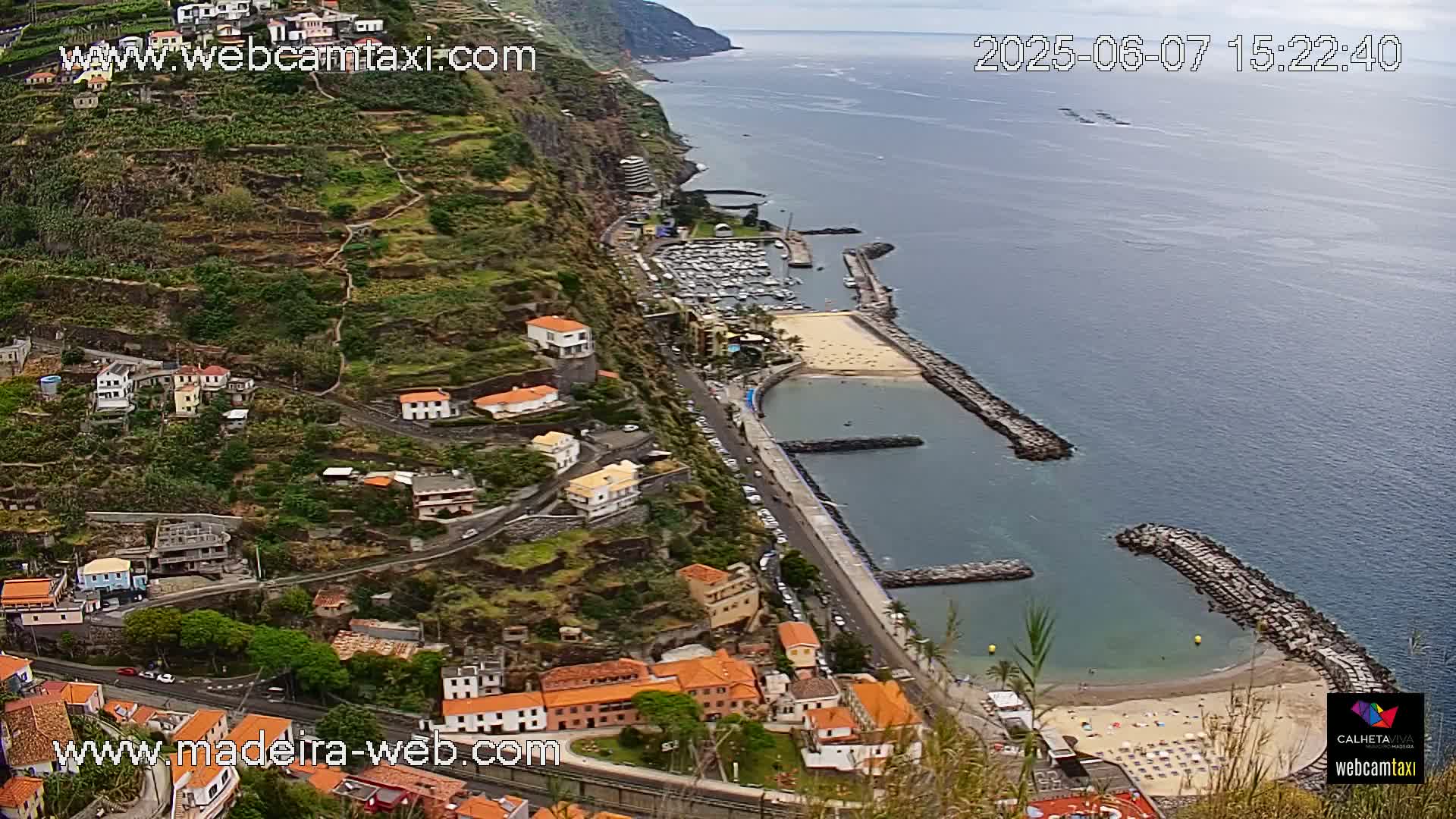 An aerial view shows a coastal town nestled on a steep hillside overlooking a calm ocean with a small marina and sandy beach.