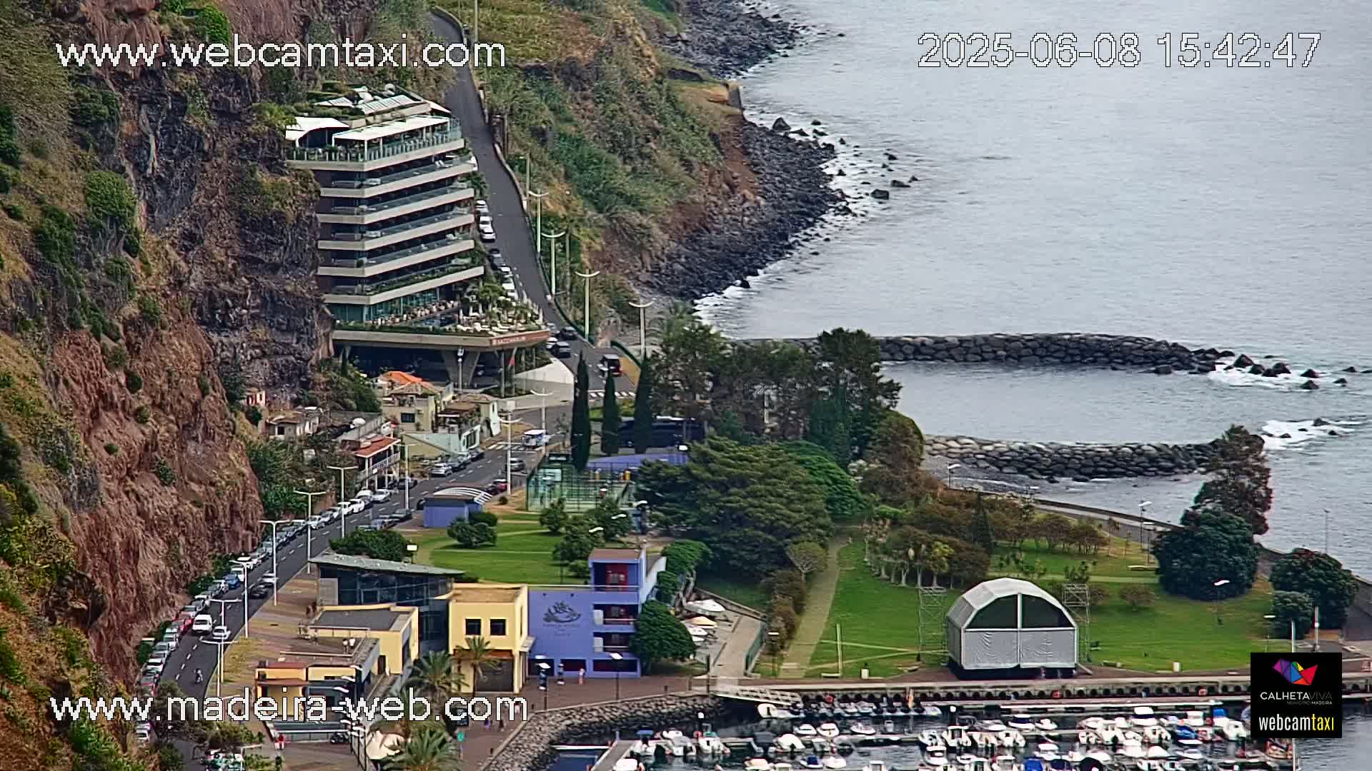 A seaside resort town built into a cliffside, featuring a multi-story hotel, a marina filled with boats, and green parkland, under a mostly cloudy sky.