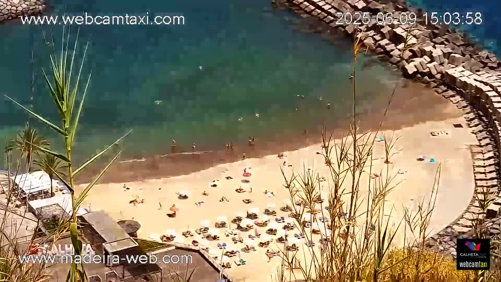 An aerial view of a sandy beach with many people sunbathing under umbrellas, partially enclosed by a rock wall, next to clear blue water on a sunny day.