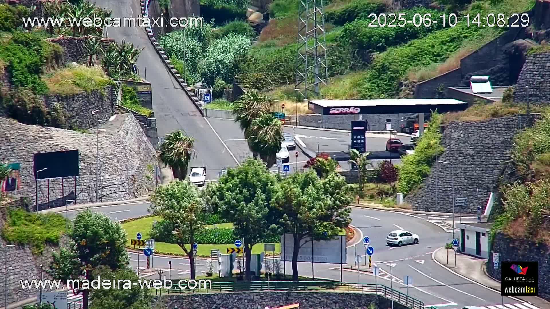 A sunny day overlooks a roundabout and a gas station nestled between steep, stone-walled hillsides, with several cars visible on the roads.