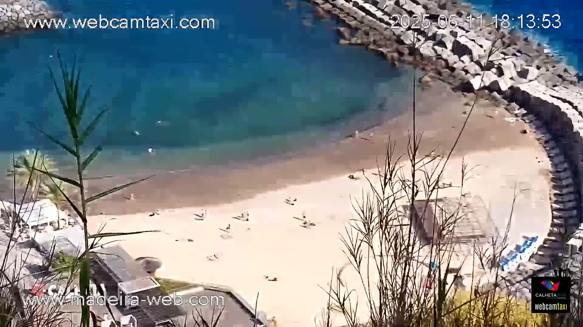 An aerial view shows a sandy beach curving into a clear blue ocean, partially enclosed by a rock wall, with several people visible on the beach and in the water on a sunny day.