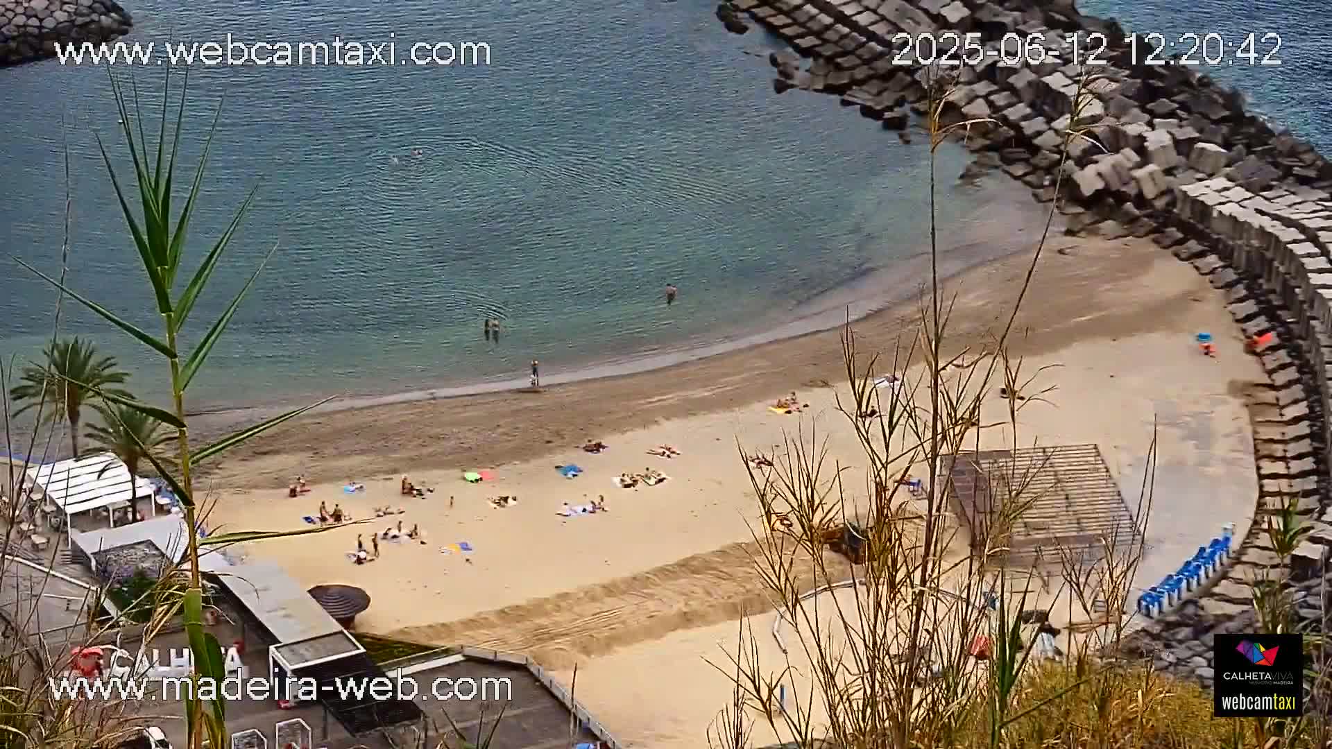 A sandy beach with scattered sunbathers and swimmers in calm, clear water is partially enclosed by a stone seawall, viewed from an elevated vantage point.