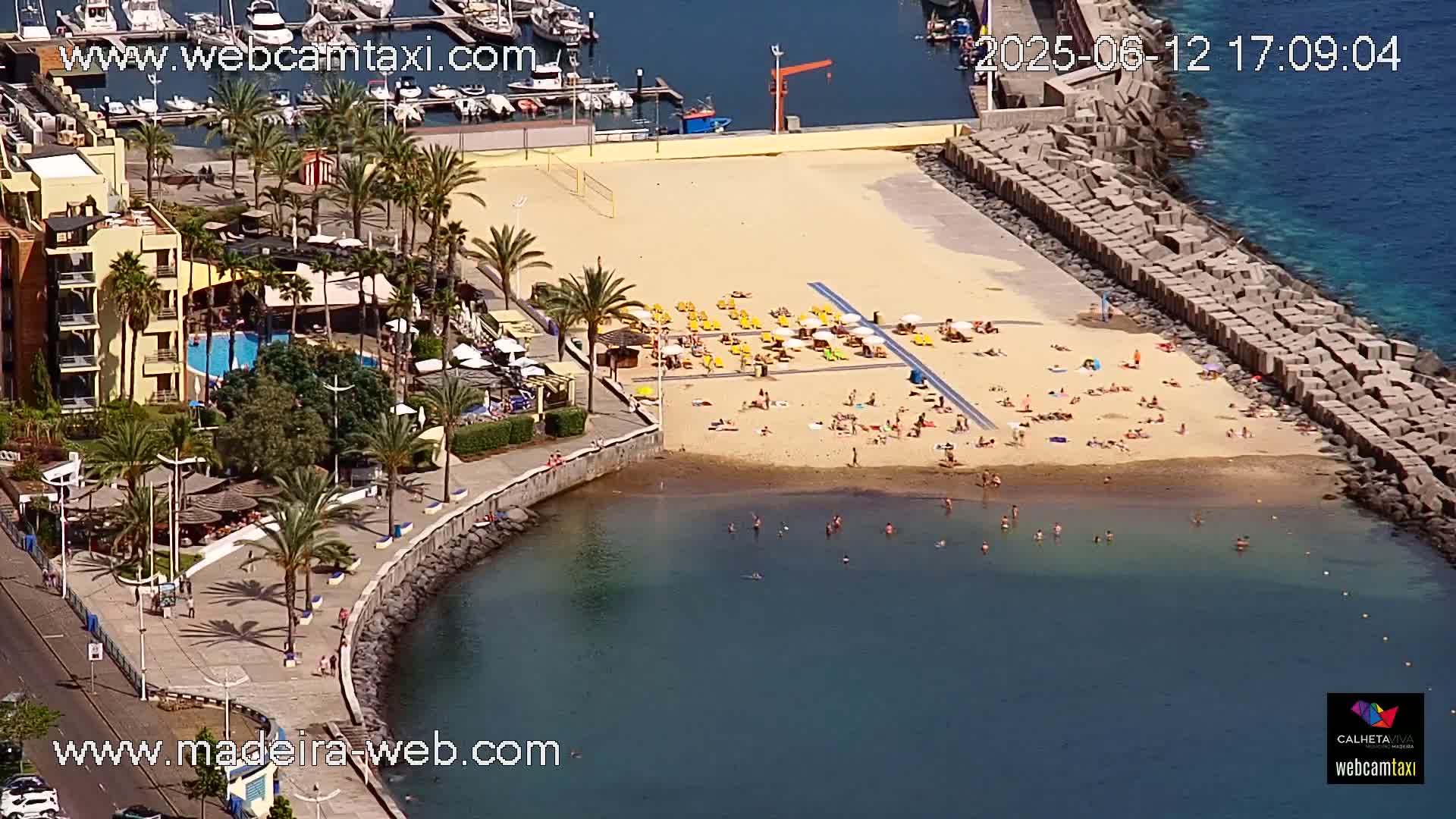 An aerial view shows a sandy beach with many sunbathers and swimmers, adjacent to a resort with a pool and marina, on a sunny day.