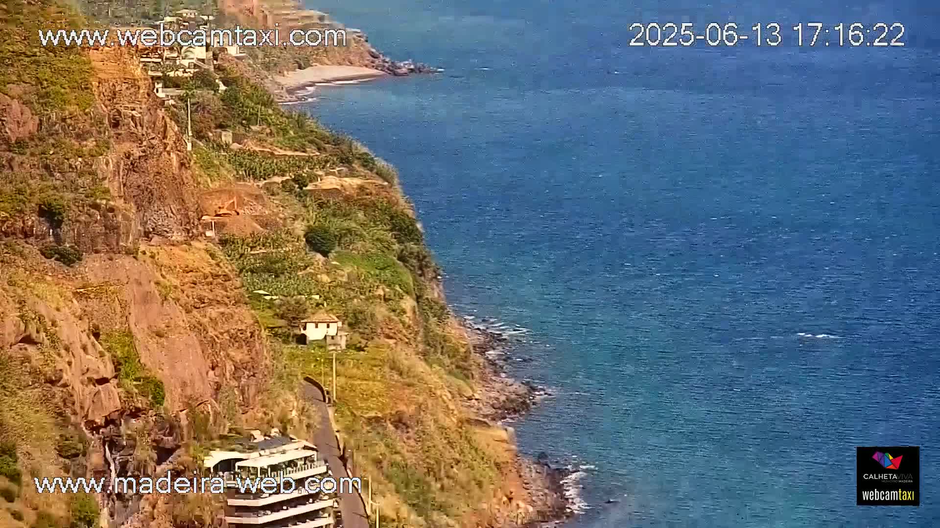 A seaside cliff with vegetation and a building overlooks a calm blue ocean on a sunny day.