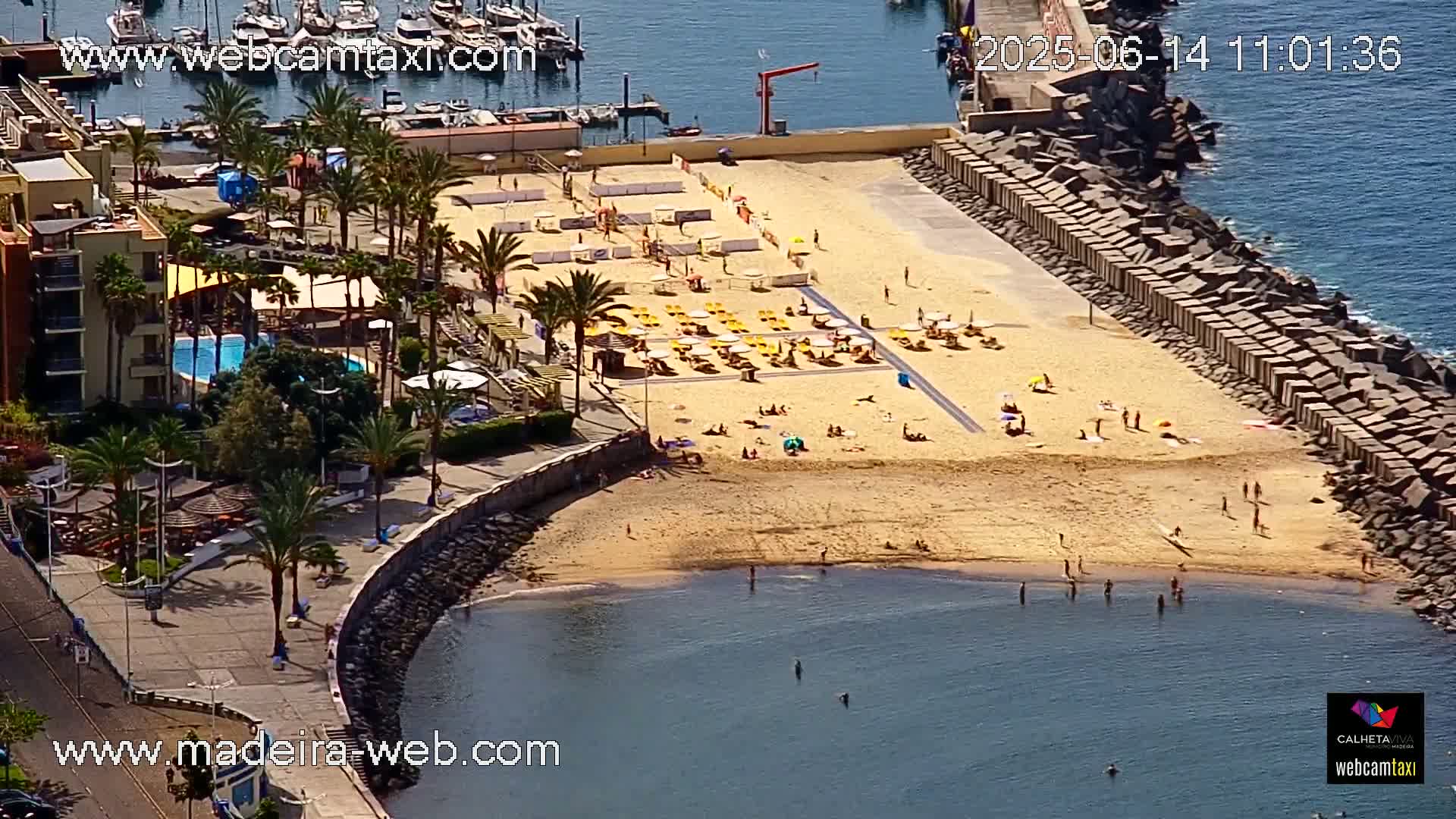 A sandy beach with people sunbathing and swimming, surrounded by a harbor with boats and a building with a pool, all under sunny skies.