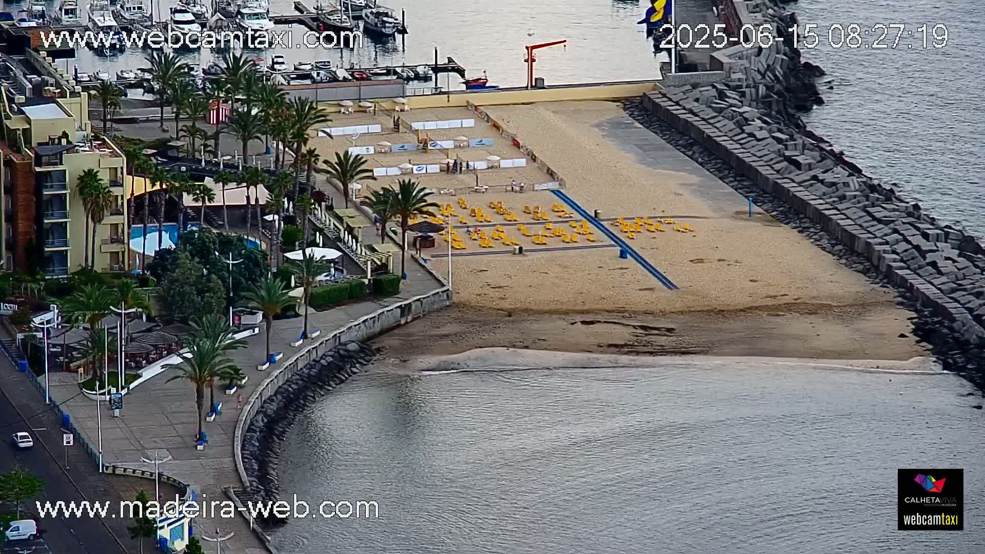 A sandy beach with yellow lounge chairs, a boardwalk lined with palm trees, a building with a pool, and a harbor in the background under partly cloudy skies.