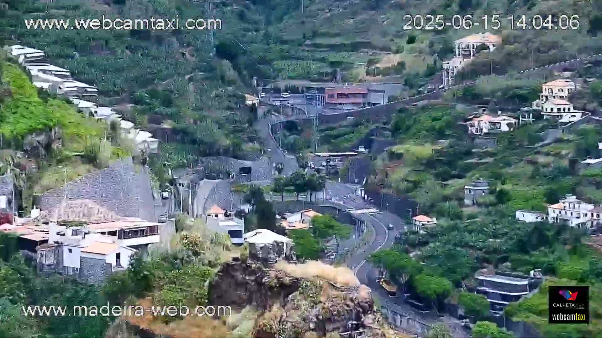A high-angle view shows a winding road through a lush, mountainous village with buildings nestled into the hillside on a seemingly clear day.