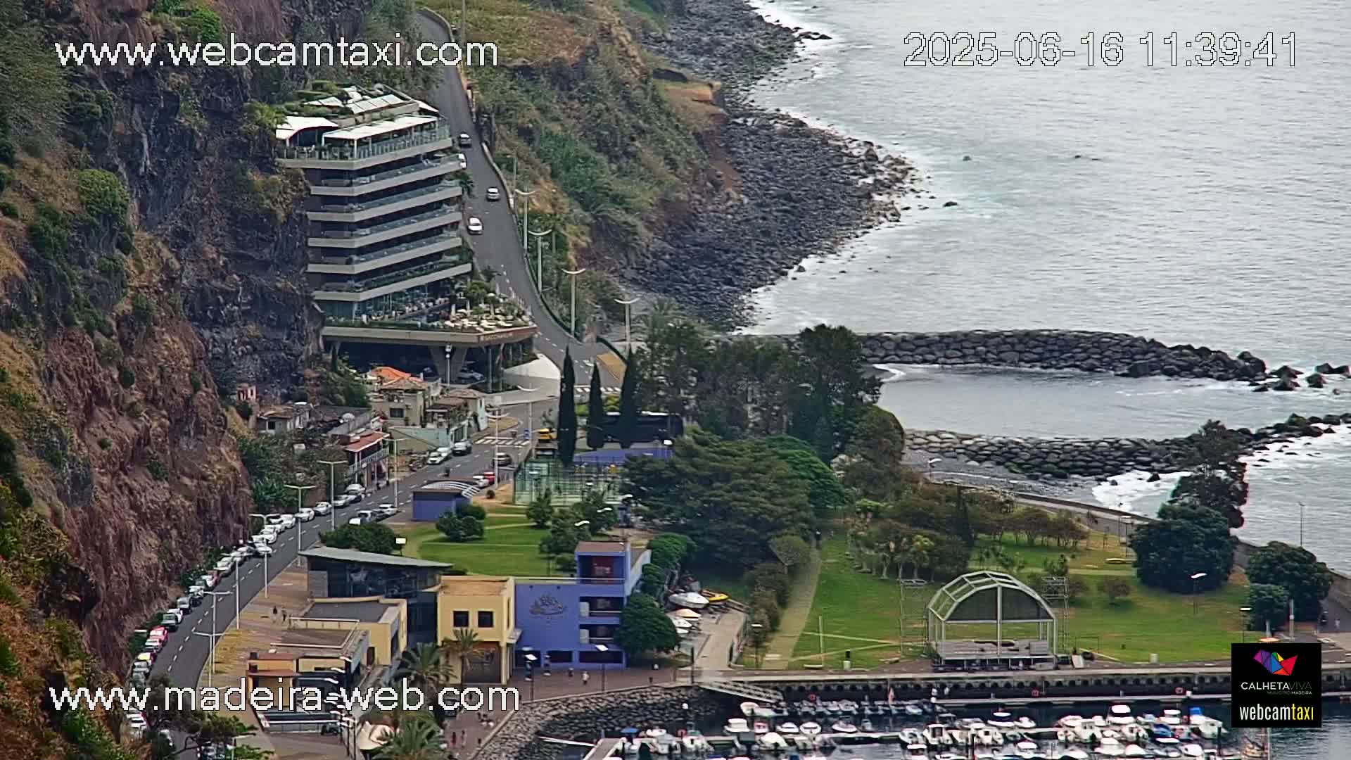 An aerial view shows a coastal town nestled against a steep, rocky cliffside next to a calm ocean, with a marina and several buildings visible.
