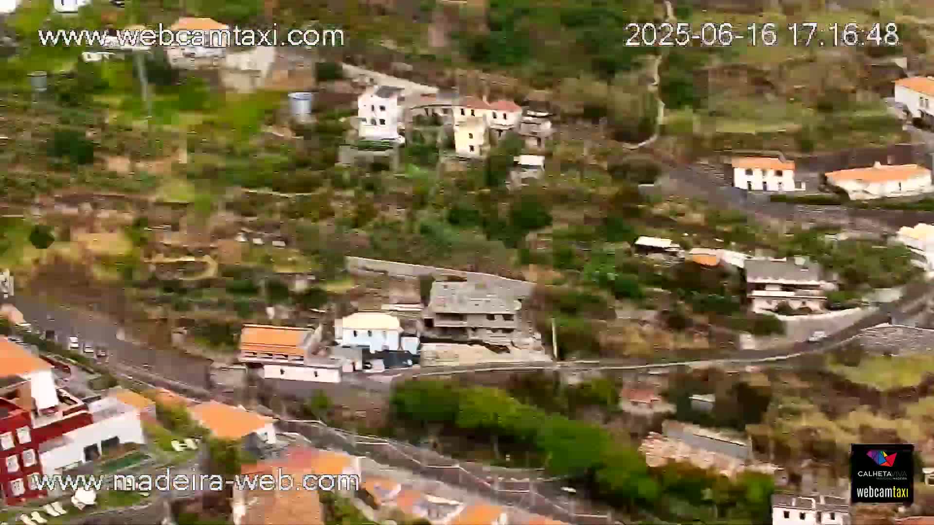 An aerial view shows a hillside village with numerous houses and roads winding through lush green vegetation under sunny skies.