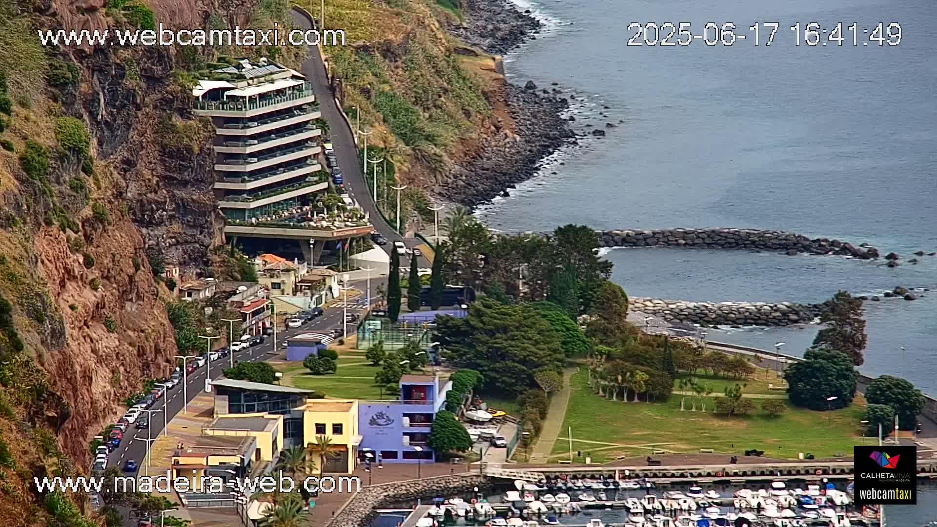 An aerial view shows a multi-story hotel built into a cliff overlooking a calm ocean with a small harbor filled with boats and a grassy park area.