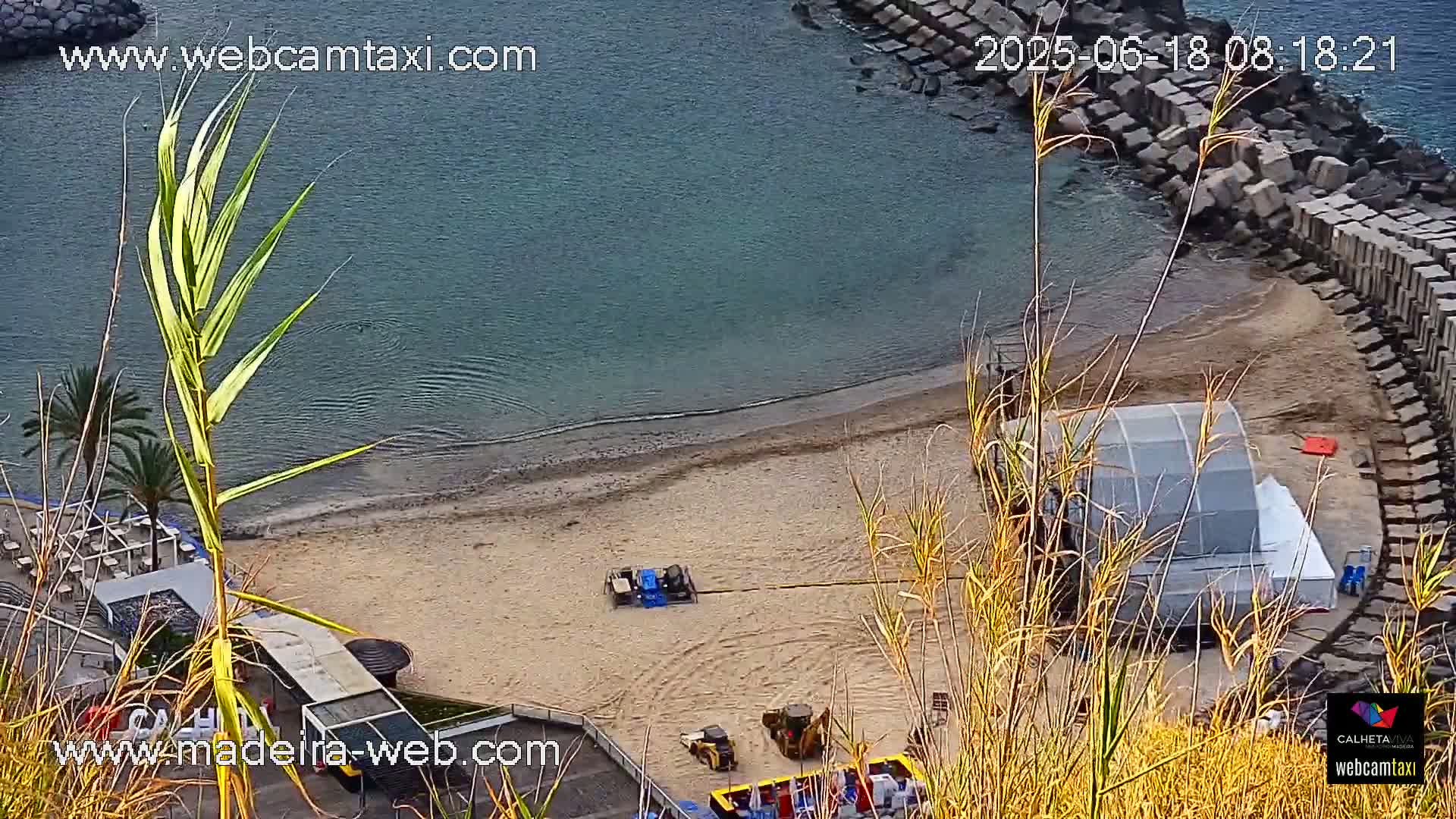 An aerial view shows a sandy beach next to calm ocean water, a large temporary structure on the sand, and construction equipment near a stone seawall.