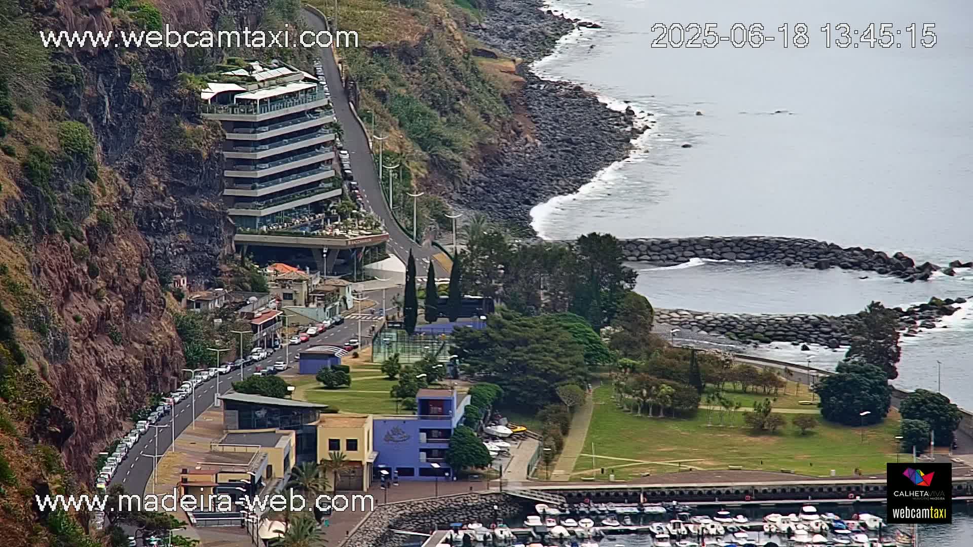 A coastal scene shows a multi-story building on a cliff overlooking a calm ocean with a small harbor filled with boats, a park, and a road with parked cars.