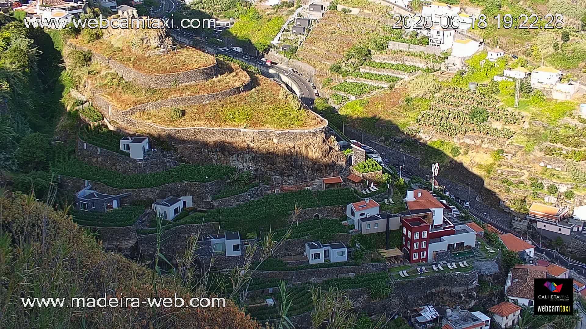 A hillside landscape with terraced farmland and modern houses, partially shaded by late afternoon sun.