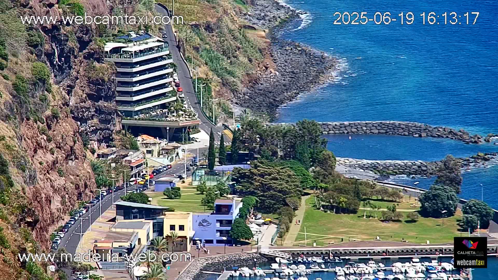 A coastal scene shows a multi-story building perched on a cliff overlooking a calm, blue ocean with a small harbor filled with boats and a grassy park area.