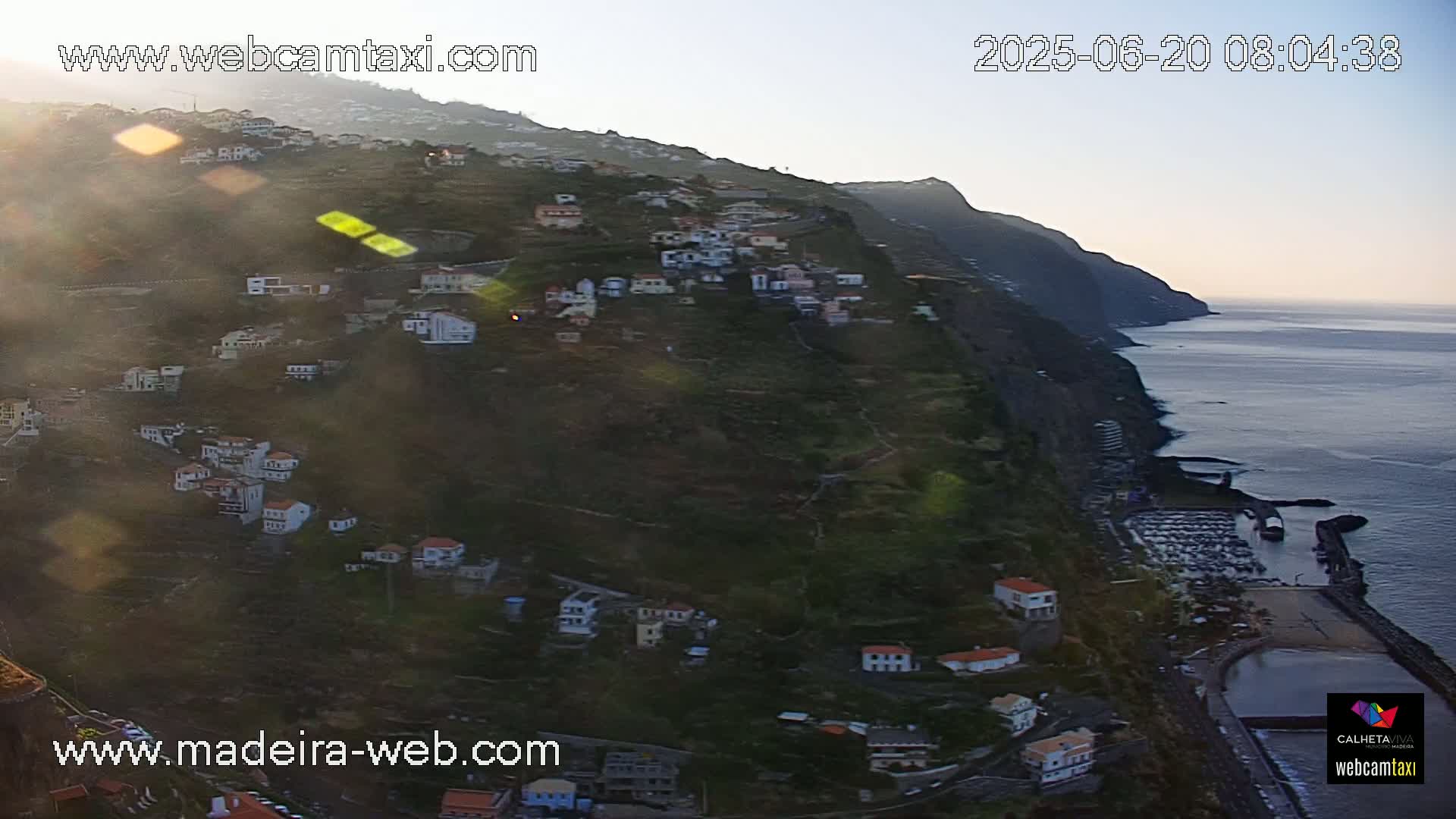 A coastal village clings to a steep, green hillside overlooking a calm ocean under a clear, bright sky.