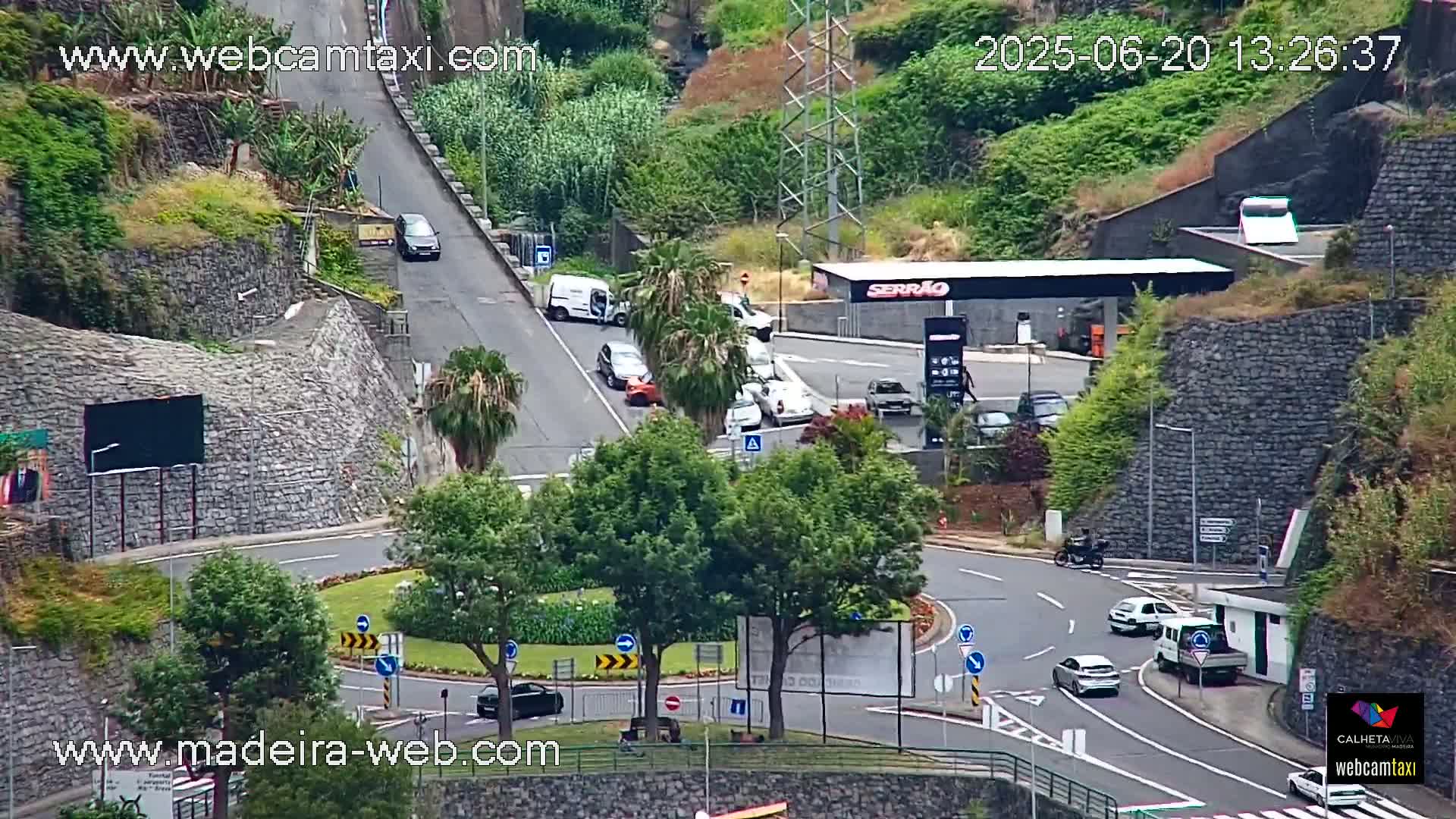 A roundabout and gas station are visible in a hilly area with lush green vegetation, under a mostly clear sky.