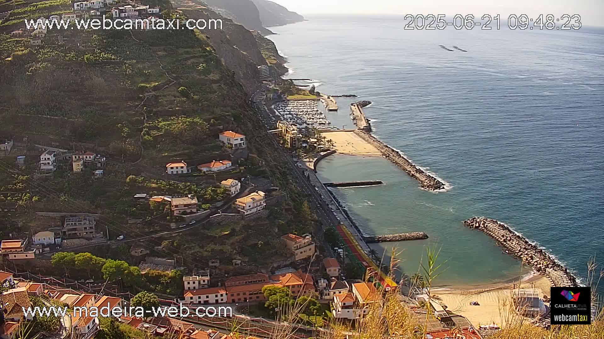 A coastal town nestled on a hillside overlooking a calm ocean with a small sandy beach and marina, on a sunny day.
