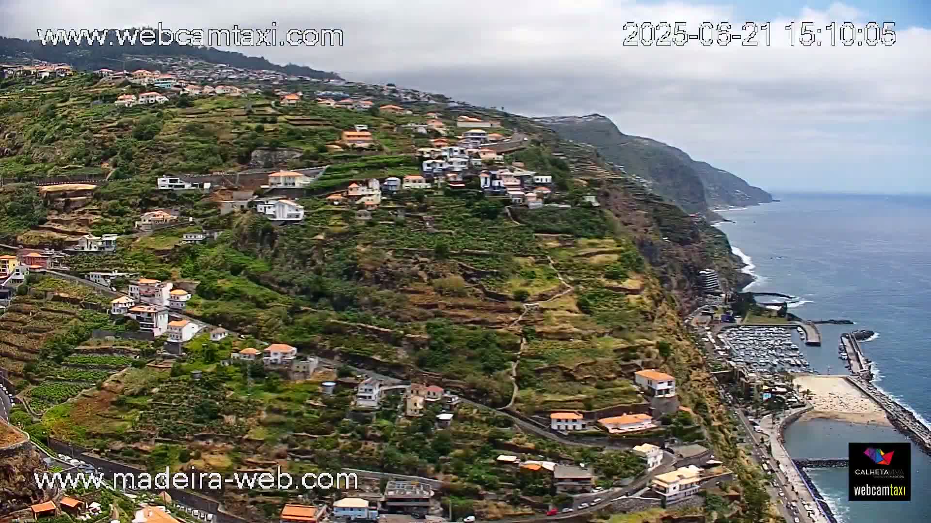 A hillside village with terraced gardens overlooks a calm ocean and a small marina under mostly cloudy skies.