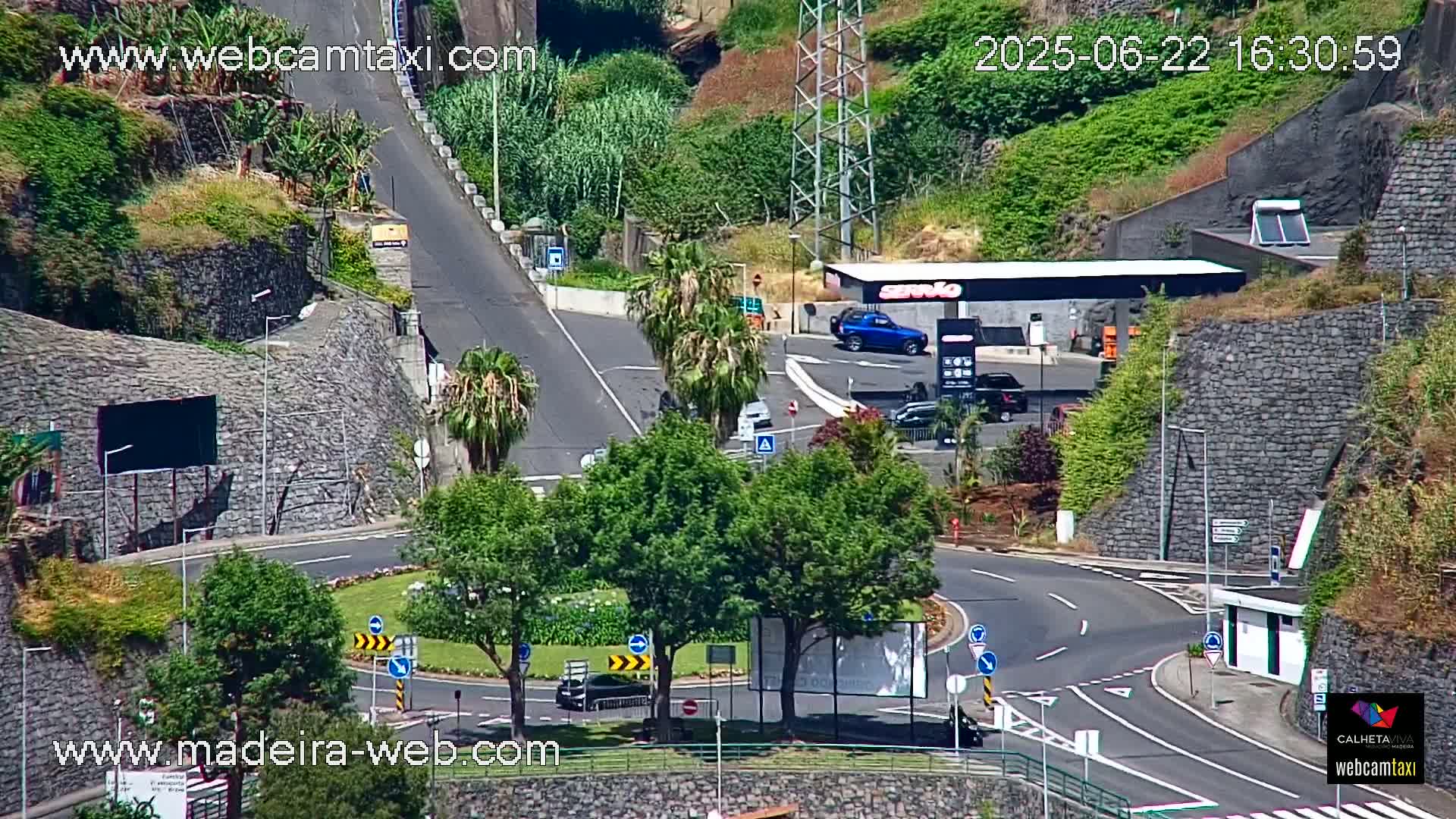 A sunny overhead view shows a roundabout and a gas station nestled between steep, green hillsides with several cars parked and driving.