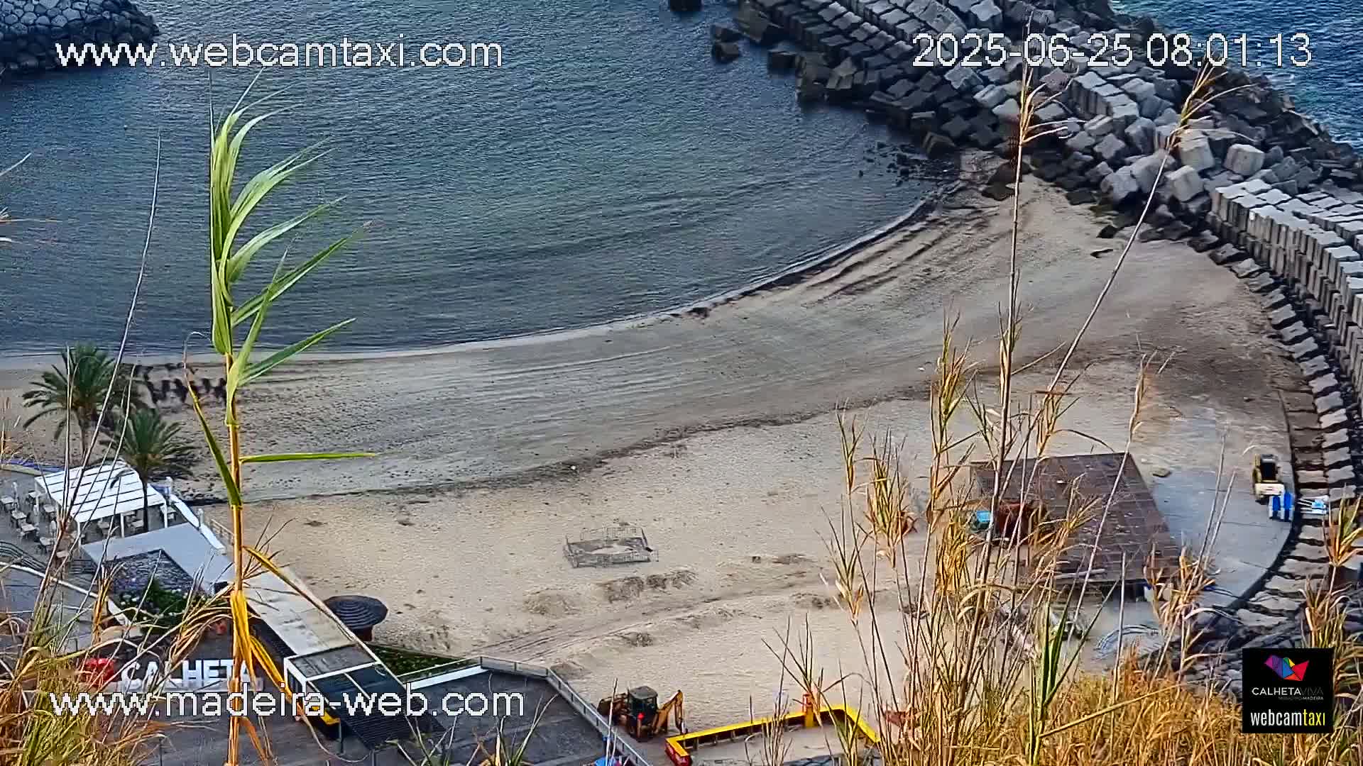 A sandy beach curves around a calm, dark-blue body of water, partially bordered by a stone wall, with some construction equipment visible near the shoreline.