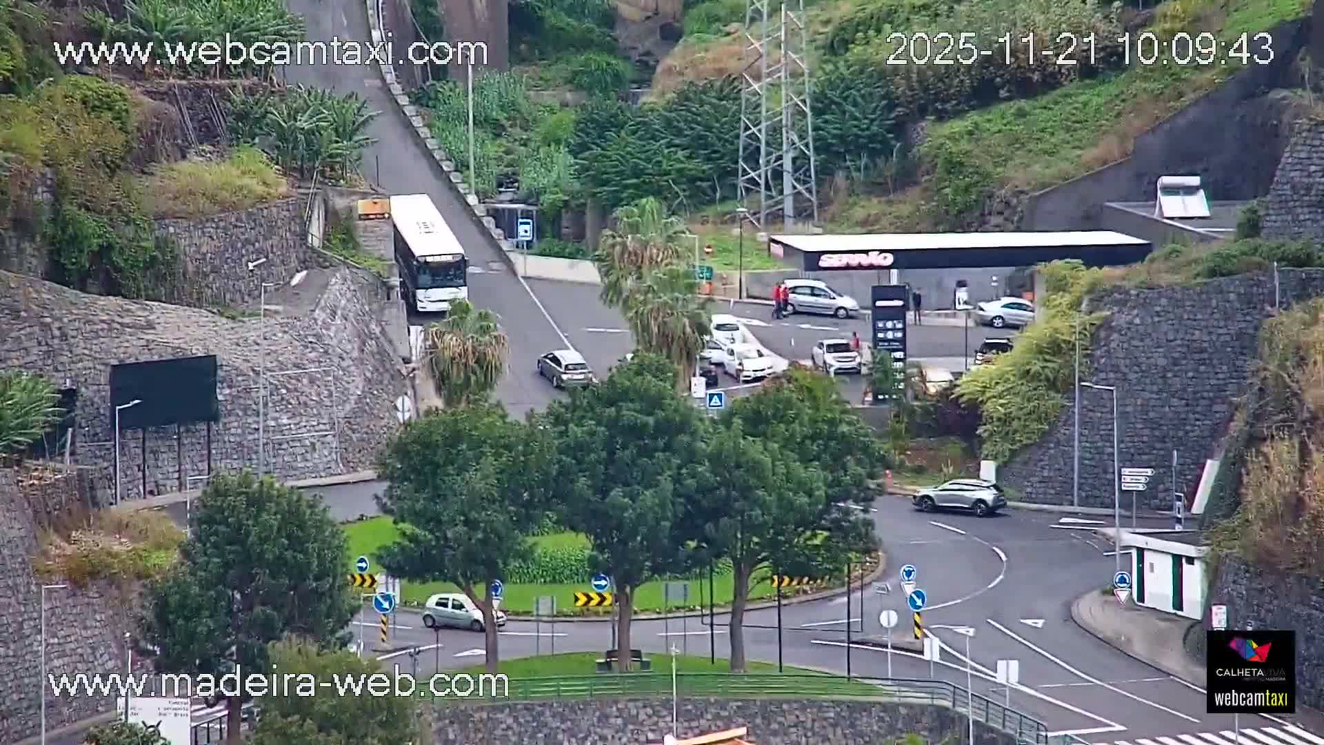 An elevated view shows a busy road network featuring a roundabout with trees, a gas station with several cars, and a white bus traveling uphill, all set amidst lush green hillsides and stone retaining walls under clear and dry daylight conditions.