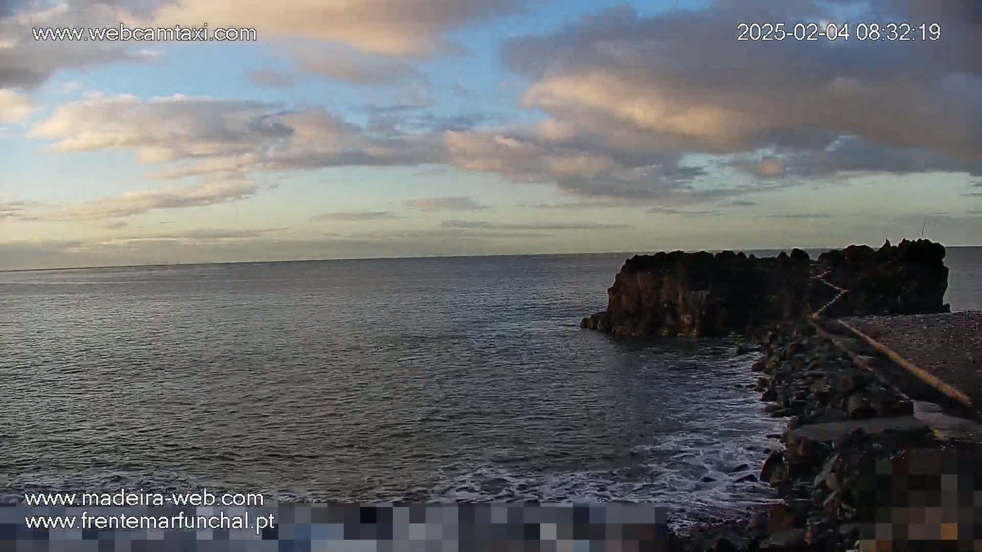 A rocky outcropping juts into a calm ocean under a partly cloudy sky.