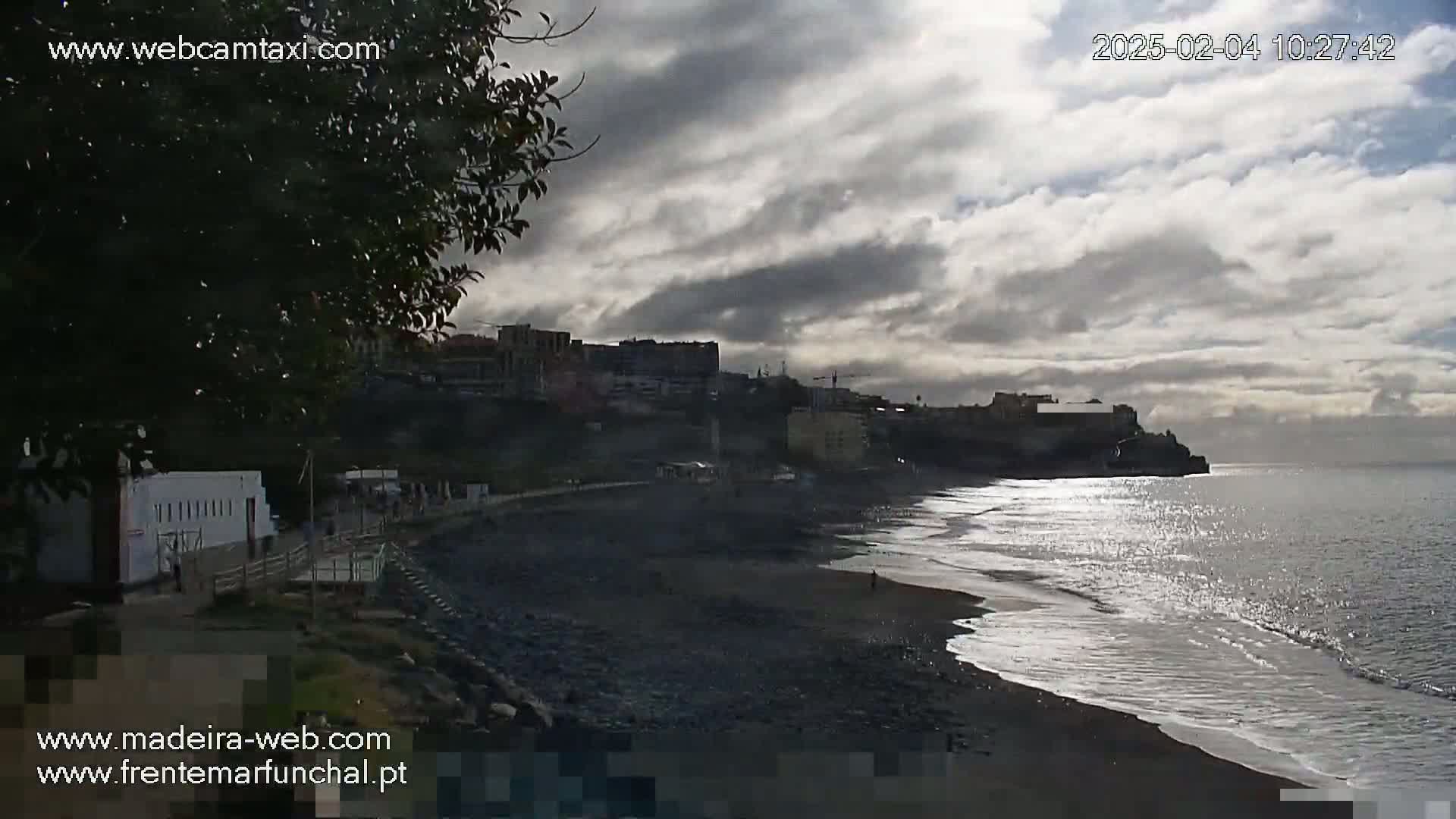 A dark-grey, cloudy sky hangs over a dark-sand beach with small waves lapping the shore, backed by a low cliff with buildings on top.