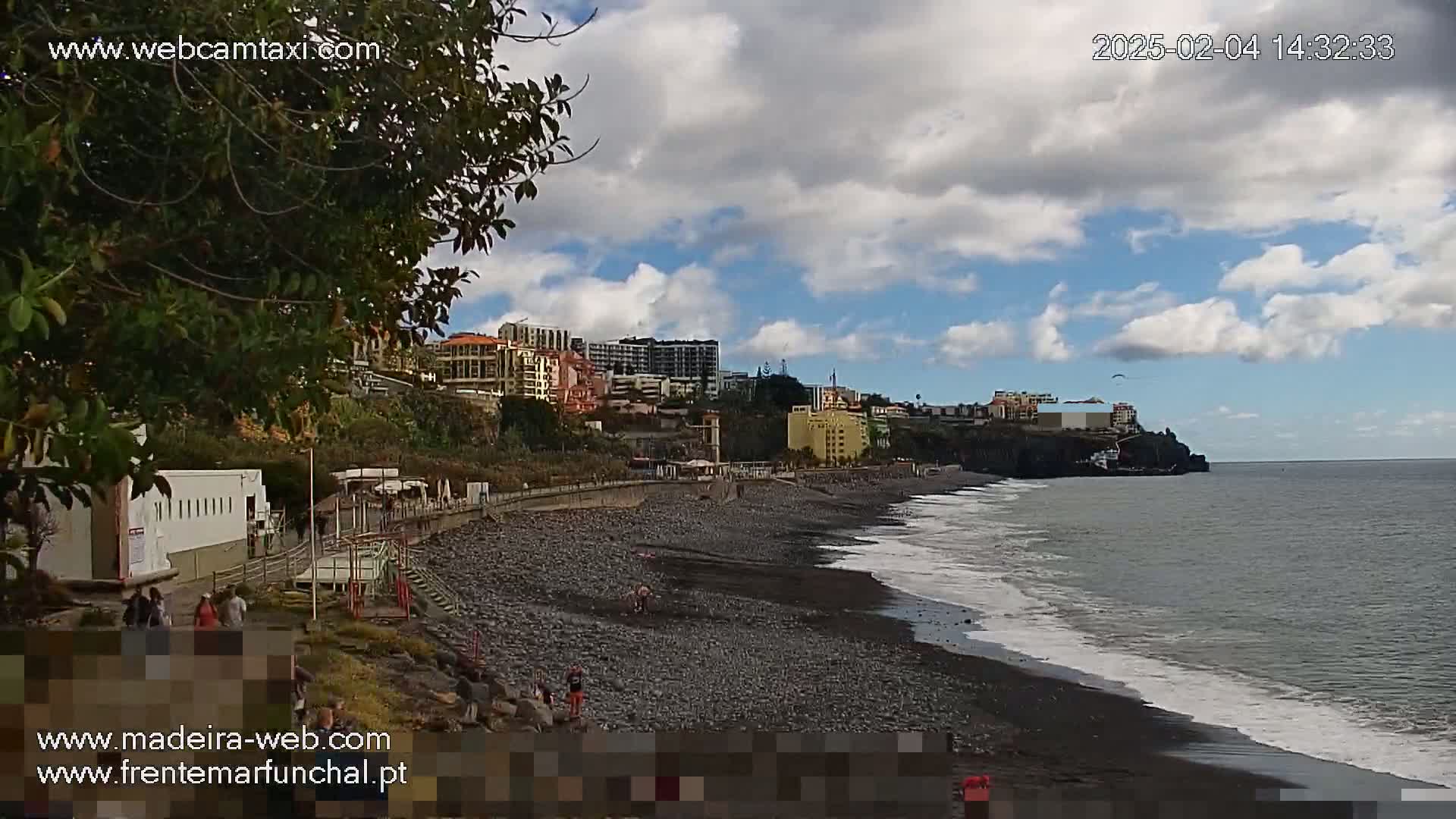 A partly cloudy day shows a dark-sand beach with a few people on it, next to a promenade and buildings that climb a hillside to meet a dark ocean.