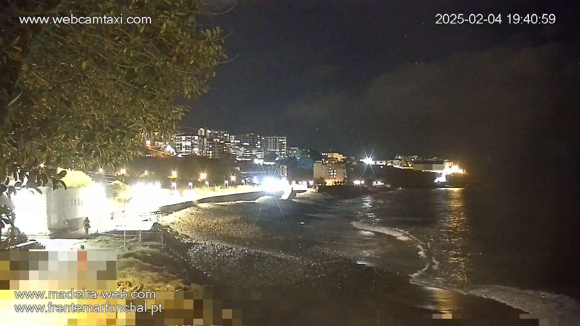 A dark, nighttime view of a pebble beach with gentle waves lapping the shore, illuminated buildings in the background, and a few lights along a walkway.
