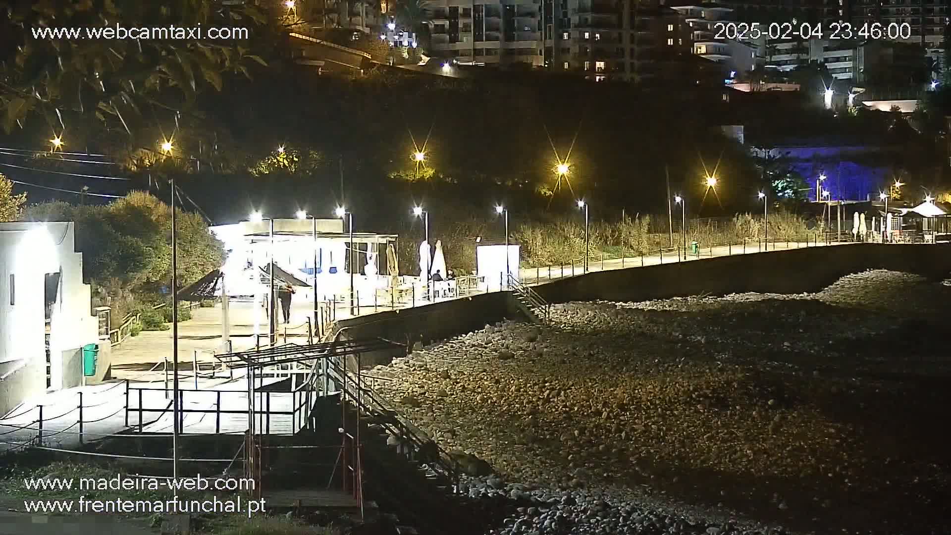 A nighttime view of a rocky shoreline with a paved walkway, outdoor seating areas, and buildings illuminated by streetlights.