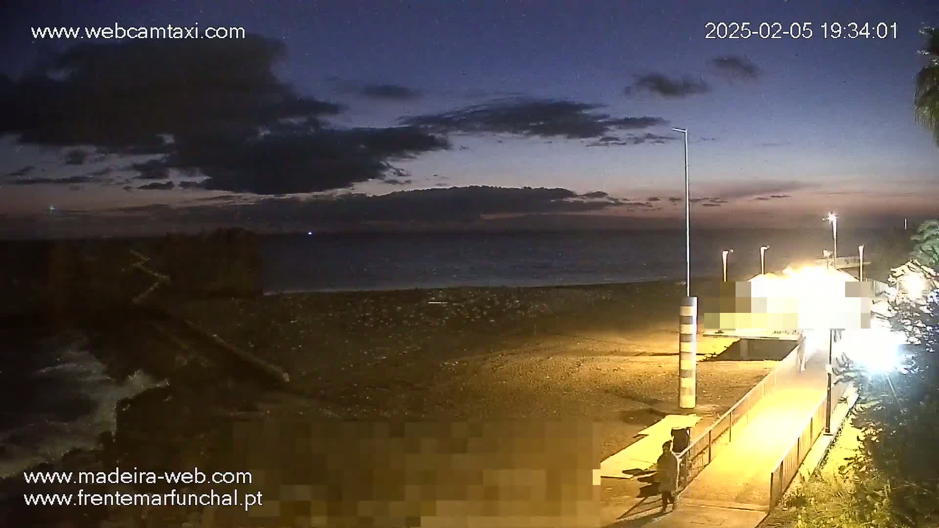 A dark, pebble beach is shown at twilight under a partly cloudy sky, with a pier and a person walking along a walkway toward the viewer.